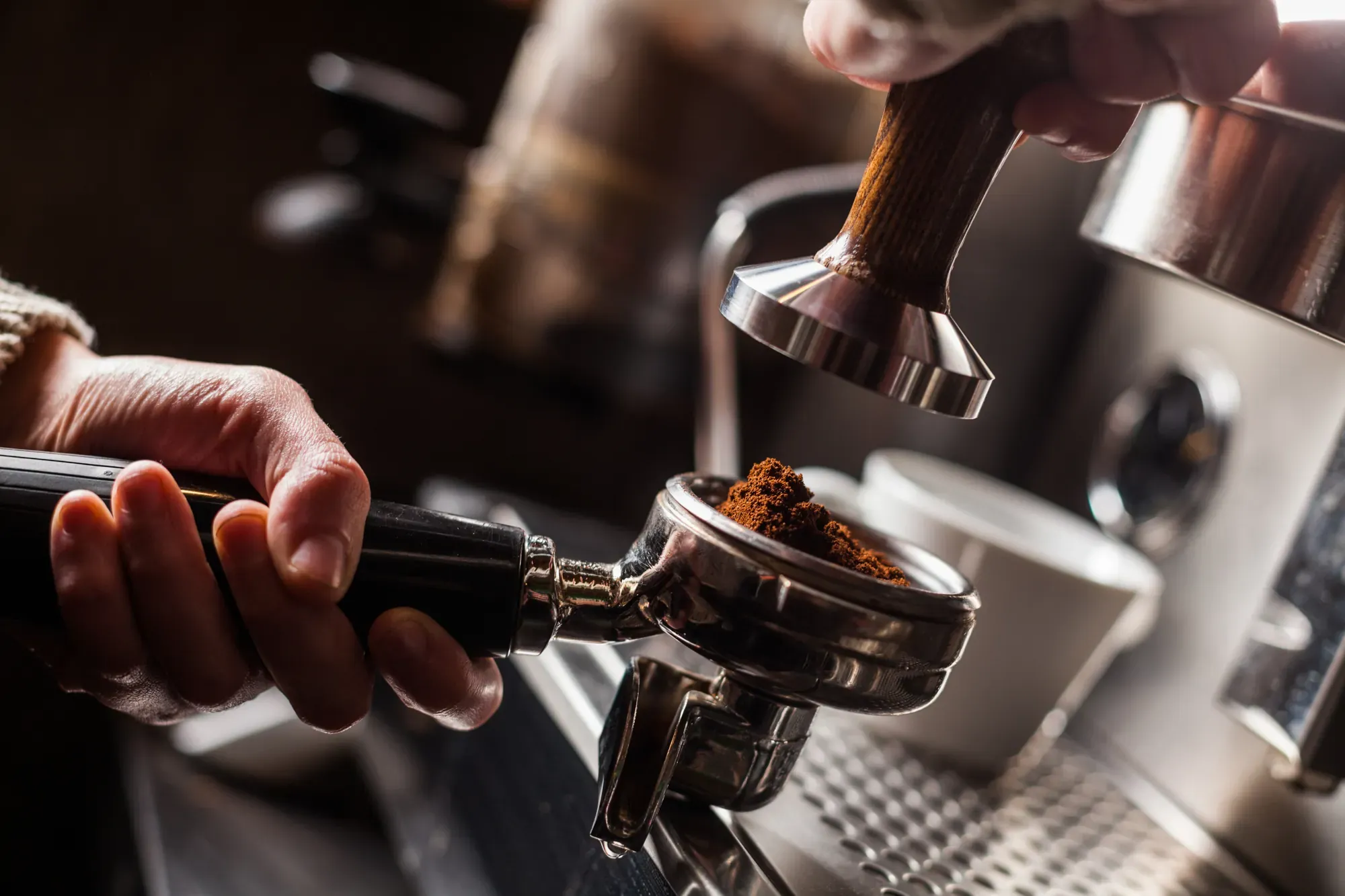 Eine Hand hält den Siebträger mit frisch gemahlenem Kaffee an der Espressomaschine, darüber der Tamper.