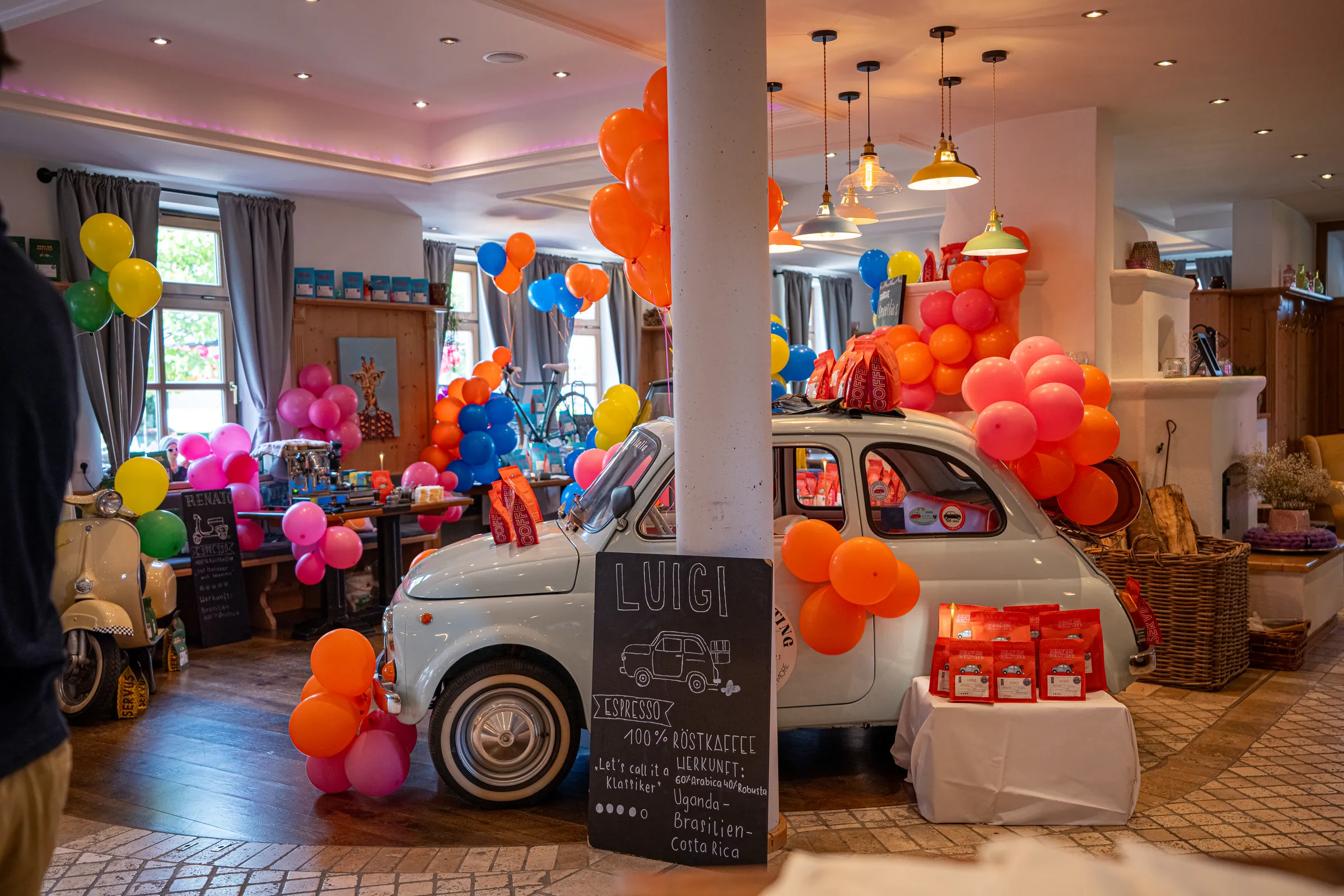 Luigis weißer Fiat 500 beim Rösterei Opening im Café, umringt von orangen Luftballons, davor die Tafel mit seinem Namen.