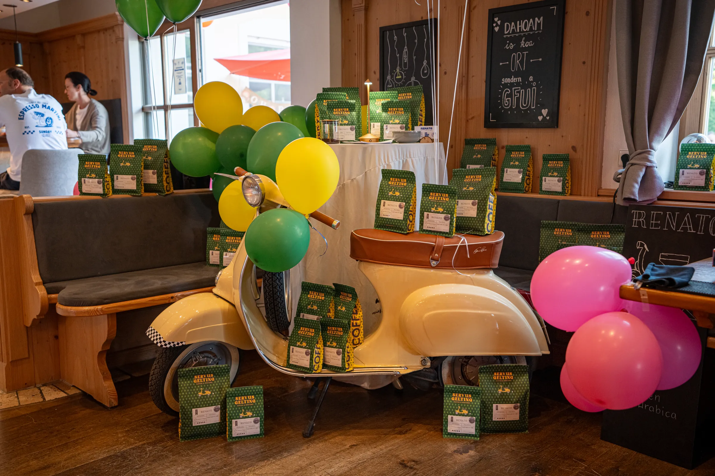 Renatos cremefarbene Vespa beim Rösterei Opening, beladen mit grünen Kaffeepackungen, dahinter Luftballons.