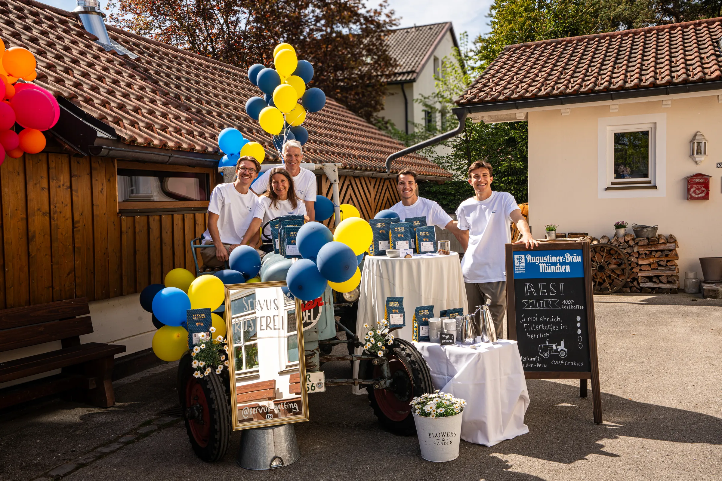 Familie und Team in weißen Servus-Gelting-Shirts stehen am alten Traktor vor der Rösterei, dazwischen Luftballons, Kaffeepackungen und die Tafel zur Sorte Resi.